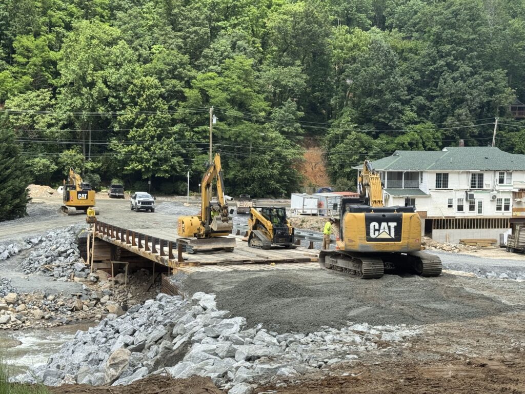 Construction crew with excavators and skid steer on temporary bridge.